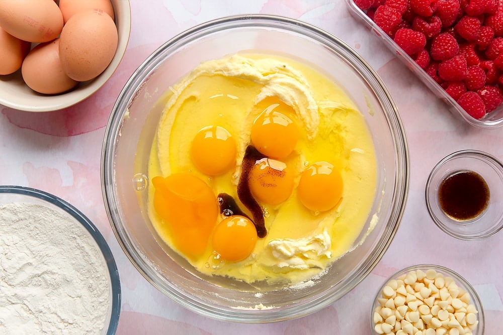 Butter and sugar beaten together in a bowl, topped with eggs and vanilla. Surrounding the bowl are ingredients to make a raspberry and white chocolate traybake