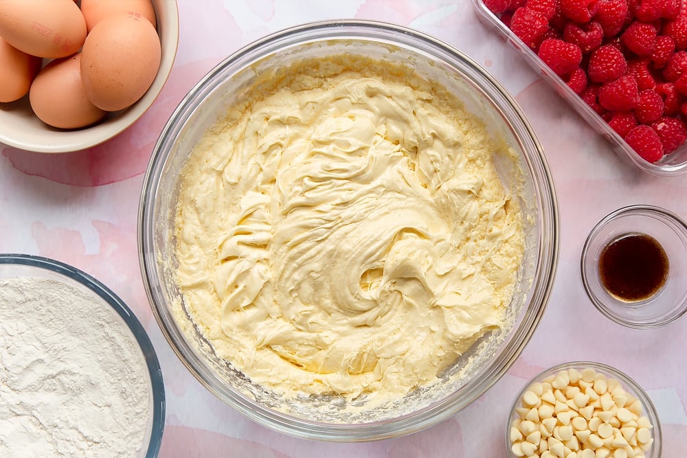 Butter, sugar, eggs and vanilla beaten together in a bowl. Surrounding the bowl are ingredients to make a raspberry and white chocolate traybake