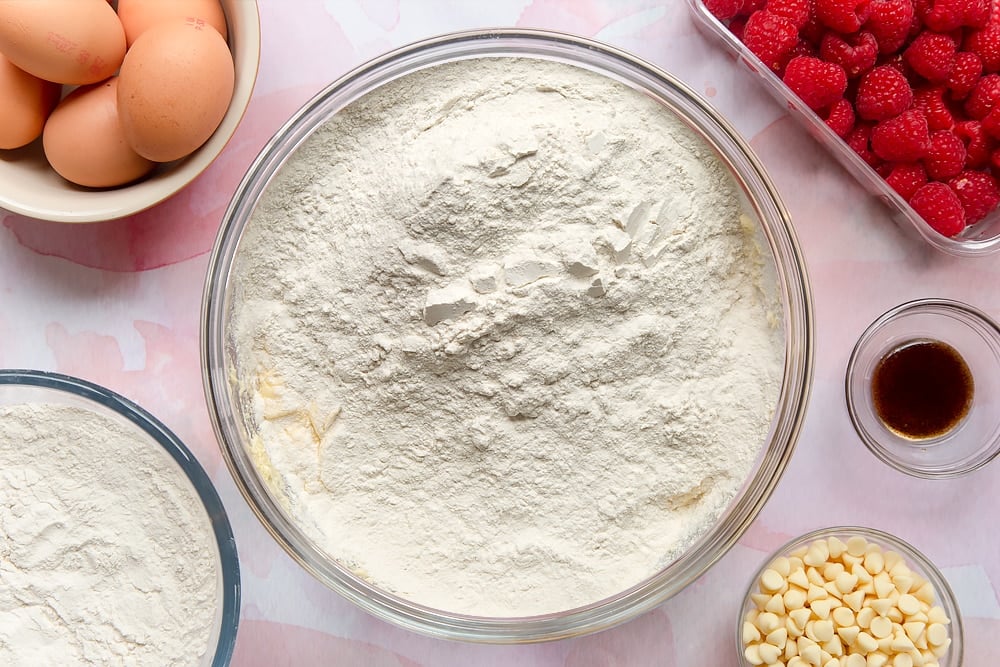 Butter, sugar, eggs and vanilla beaten together in a bowl. Flour on top. Surrounding the bowl are ingredients to make a raspberry and white chocolate traybake