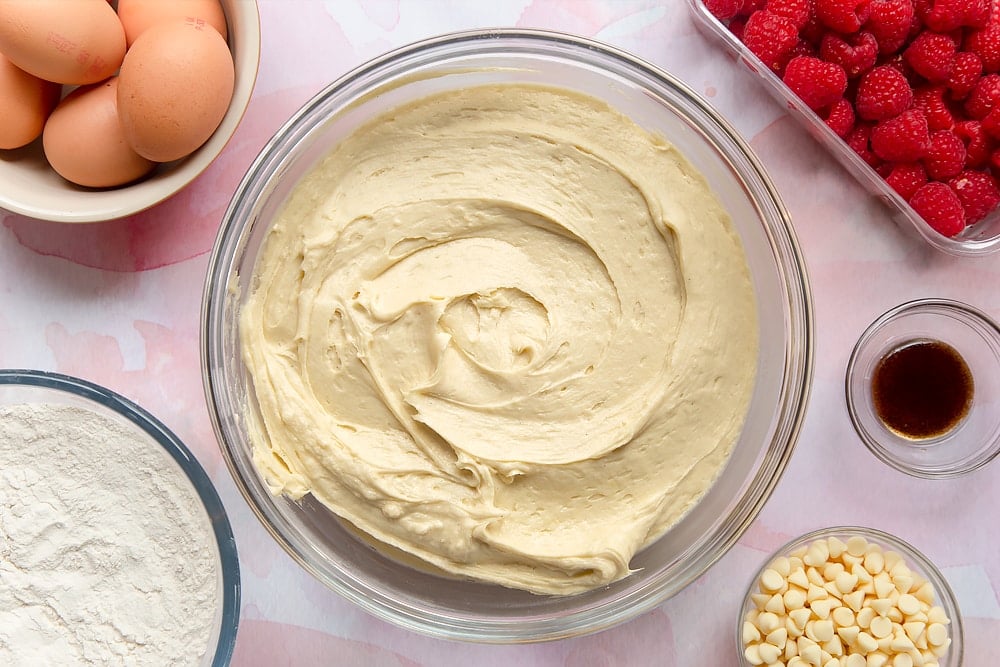 Cake batter in a bowl. Surrounding the bowl are ingredients to make a raspberry and white chocolate traybake