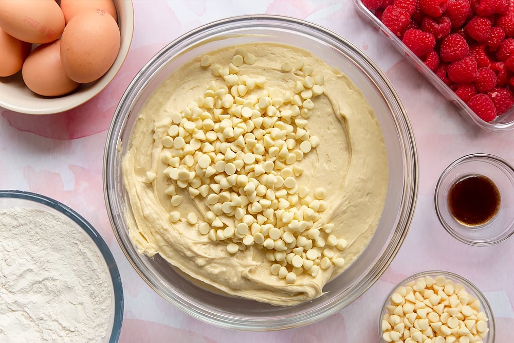 Cake batter in a bowl, topped with white chocolate chips. Surrounding the bowl are ingredients to make a raspberry and white chocolate traybake