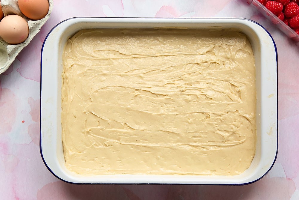 Cake batter with white chocolate chips spread in a tray. Surrounding the tray are ingredients to make a raspberry and white chocolate traybake