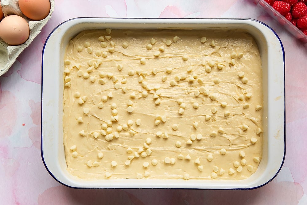 Cake batter with spread in a tray with white chocolate chips scattered on top. Surrounding the tray are ingredients to make a raspberry and white chocolate traybake