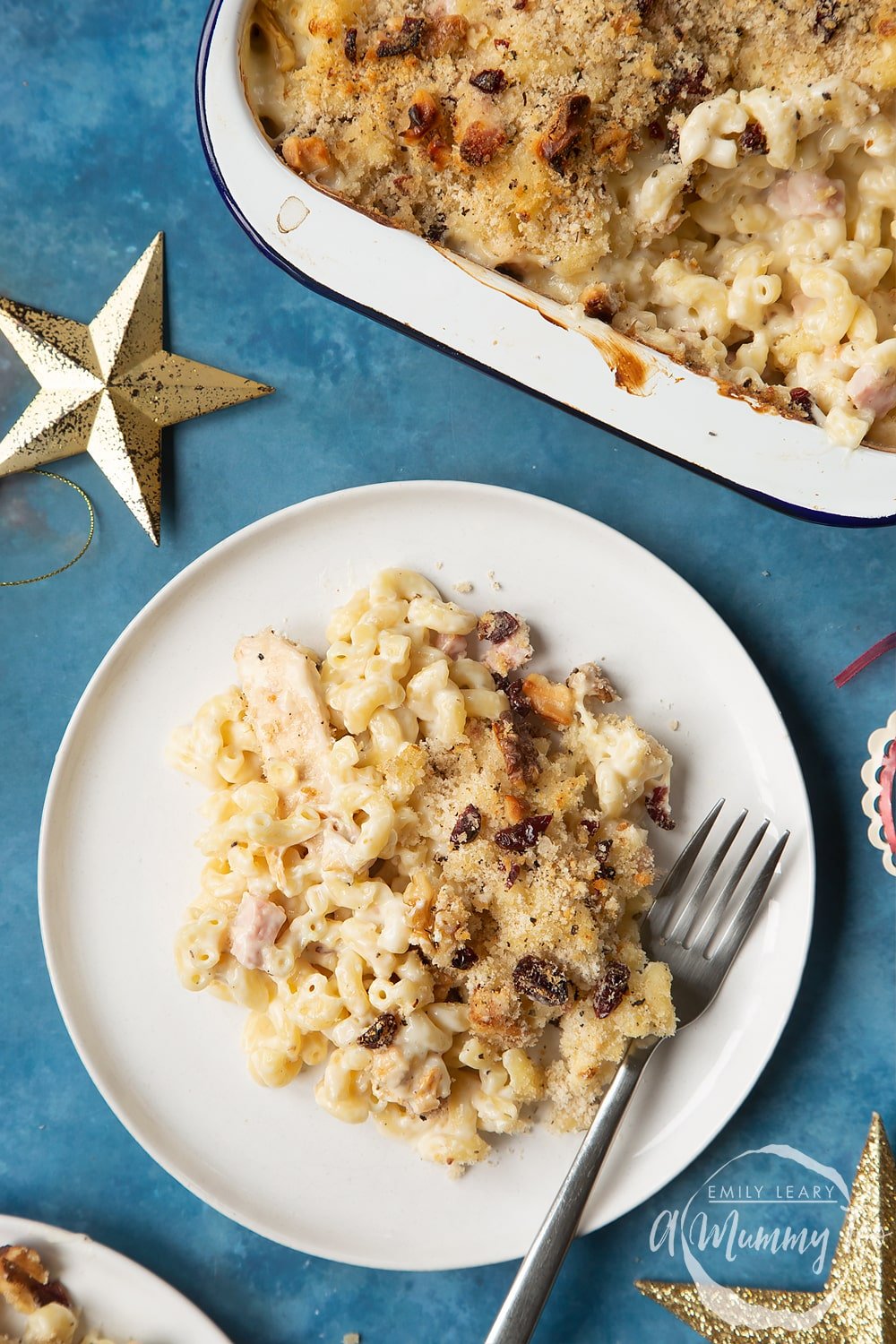 A portion of chicken and bacon mac and cheese served on a small white plate with a fork. A tray containing more of the pasta is shown to one side.