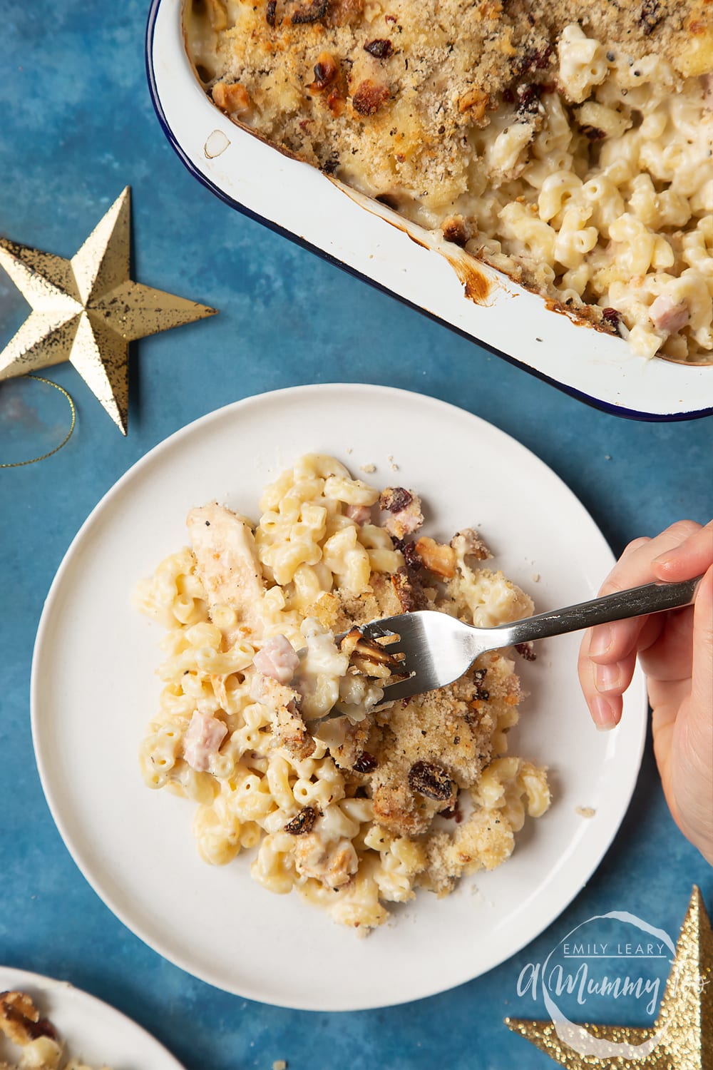 A portion of chicken and bacon mac and cheese served on a small white plate. A hand holding on a fork digs into the pasta. A tray containing more of the pasta is shown to one side.