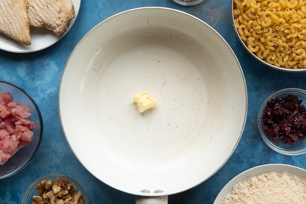 A pan containing a little bit of butter. The ingredients to make chicken and bacon mac and cheese surround the pan.