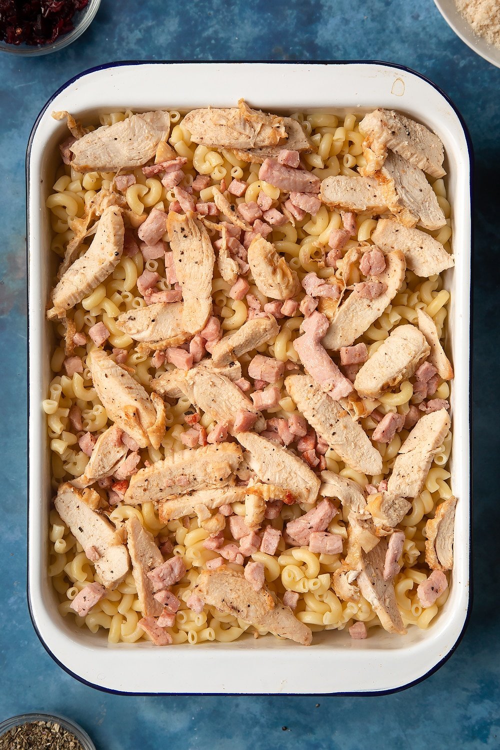 A large roasting pan filled with cooked macaroni pasta, topped with cooked chicken and bacon. The ingredients to make chicken and bacon mac and cheese surround the pan.