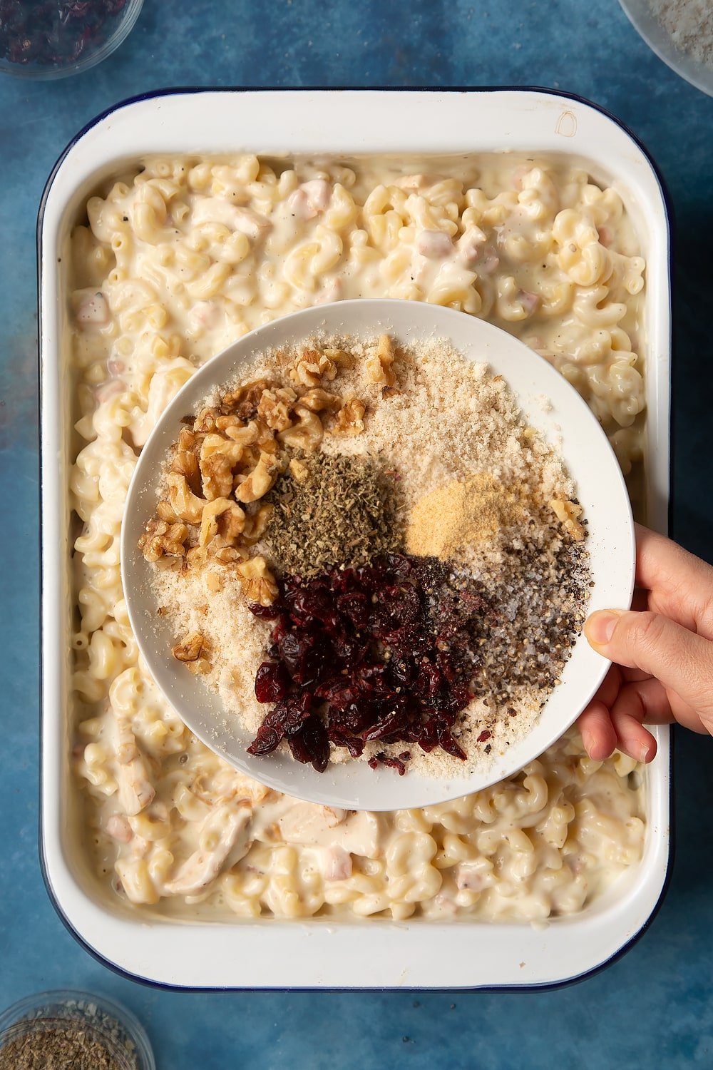 A large roasting pan filled with chicken and bacon mac and cheese. A hand holds a plate above the pan, with breadcrumbs, dried cranberries, walnuts, garlic granules, dried sage, salt and pepper.