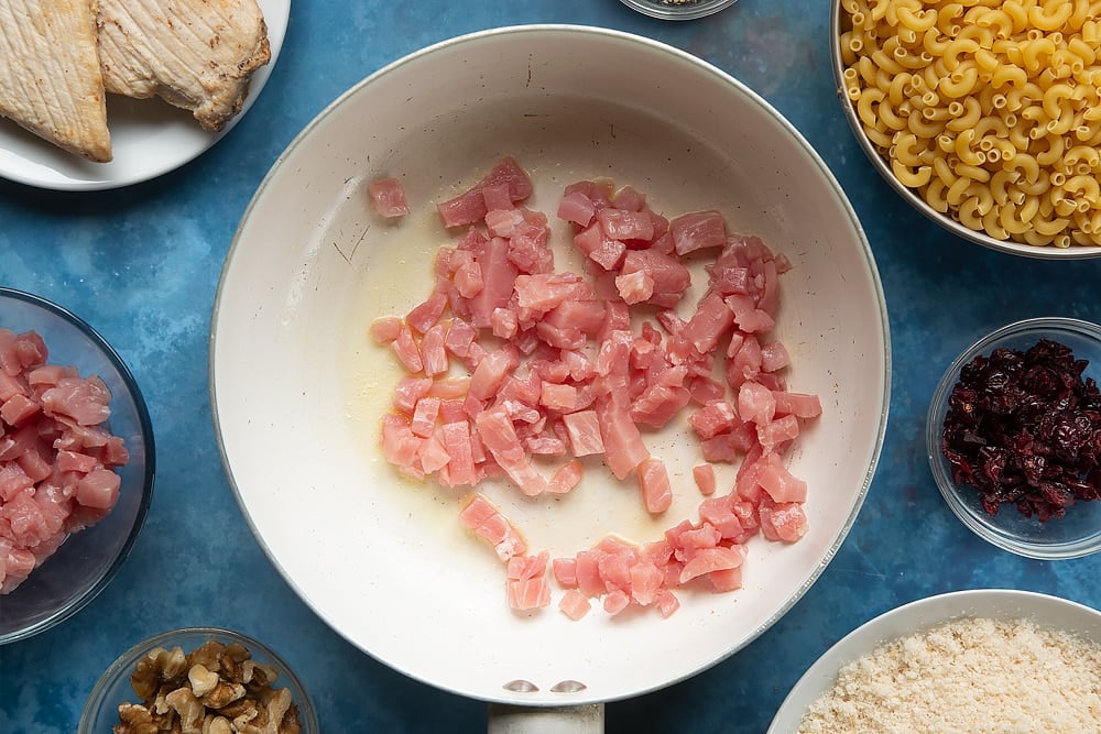 A pan containing a little melted butter and raw chopped bacon. The ingredients to make chicken and bacon mac and cheese surround the pan.