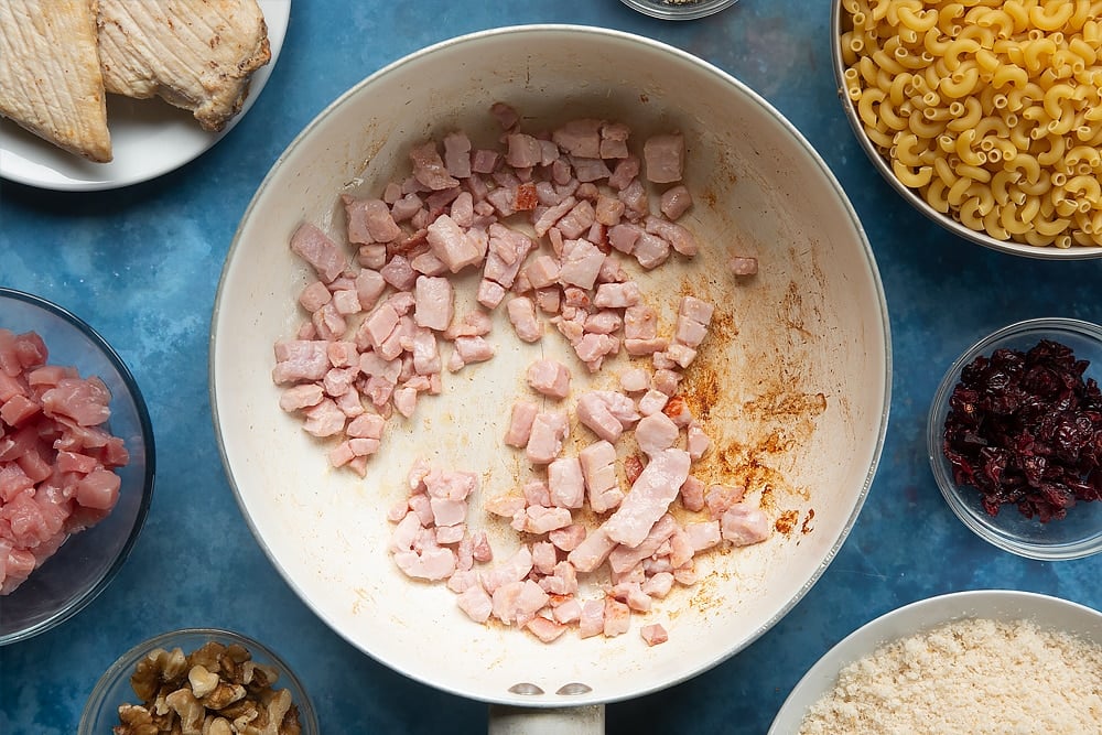 A pan containing cooked chopped bacon. The ingredients to make chicken and bacon mac and cheese surround the pan.