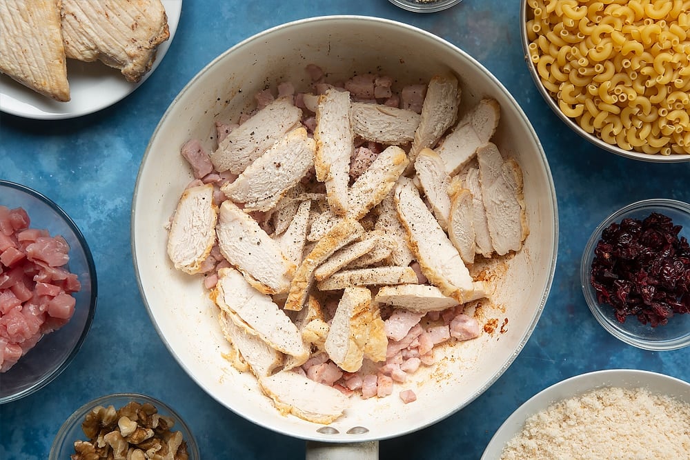 A pan containing cooked chopped bacon, cooked sliced chicken and black pepper. The ingredients to make chicken and bacon mac and cheese surround the pan.