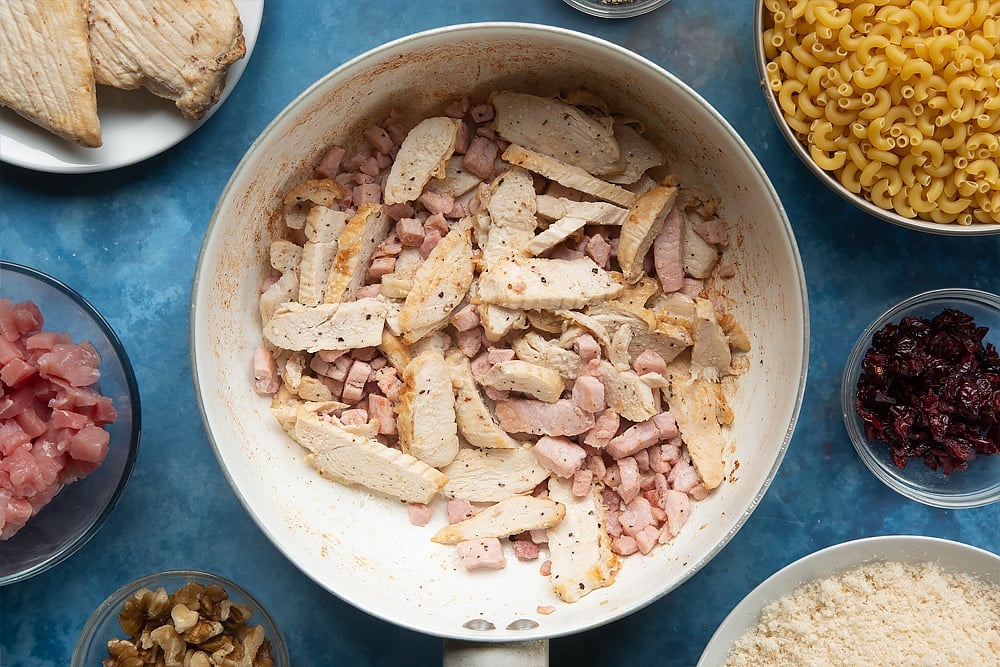 A pan containing cooked chopped bacon, chicken and black pepper, mixed together. The ingredients to make chicken and bacon mac and cheese surround the pan.