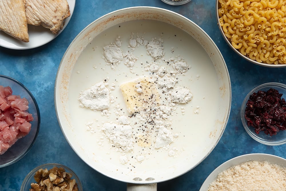 A pan containing milk, flour, butter and black pepper. The ingredients to make chicken and bacon mac and cheese surround the pan.