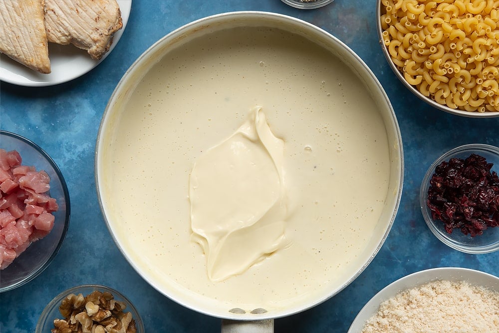 A pan containing a white sauce. Primula cheese is on top. The ingredients to make chicken and bacon mac and cheese surround the pan.