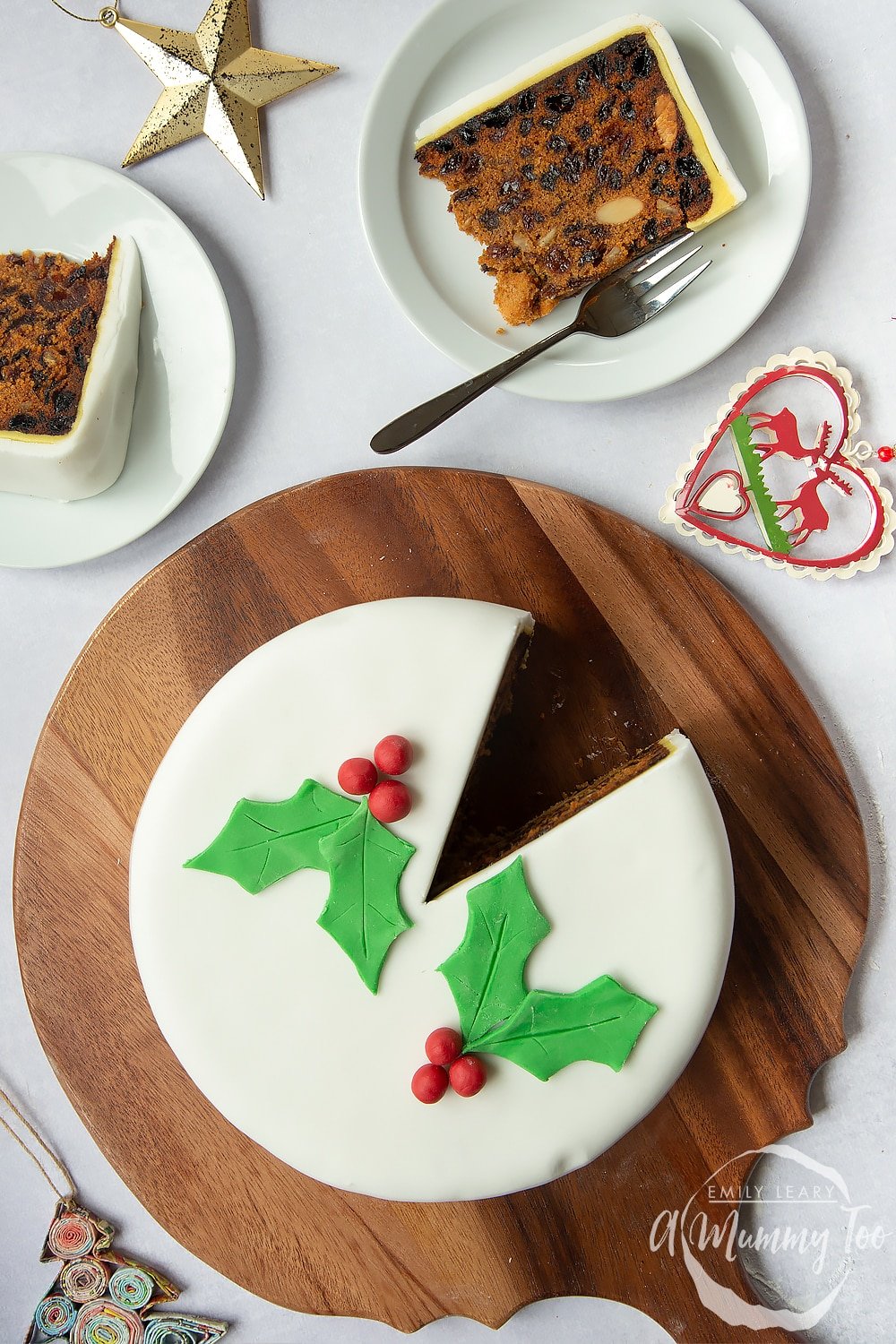 an iced christmas fruit cake on a round wooden chopping board with a slice cut out on a white plate.