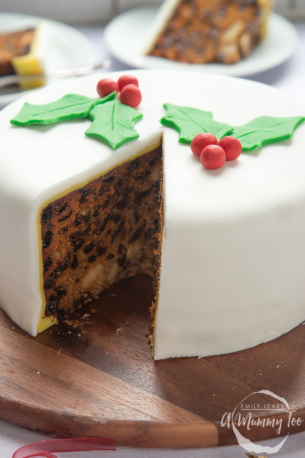 an iced christmas fruit cake on a round wooden chopping board with a slice cut out on a white plate.