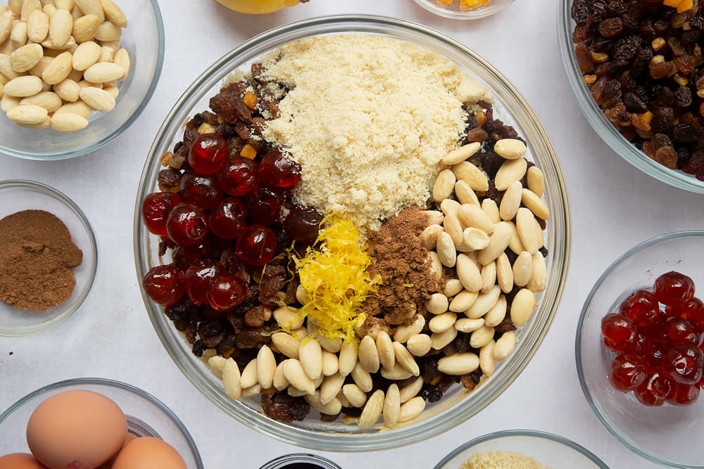 christmas fruit cake base batter mixed together in a large clear bowl topped with chopped dried fruit surrounded by ingredients in smaller clear bowls.