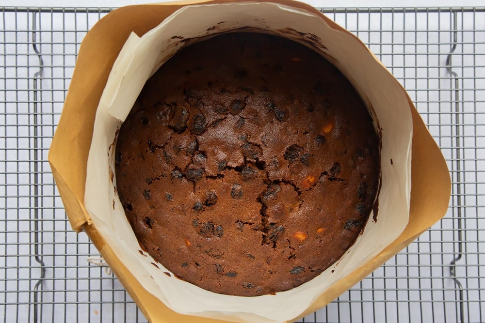 baked christmas fruit cake in a cake tin wrapped in greaseproof paper and brown paper on a wire cooling rack.