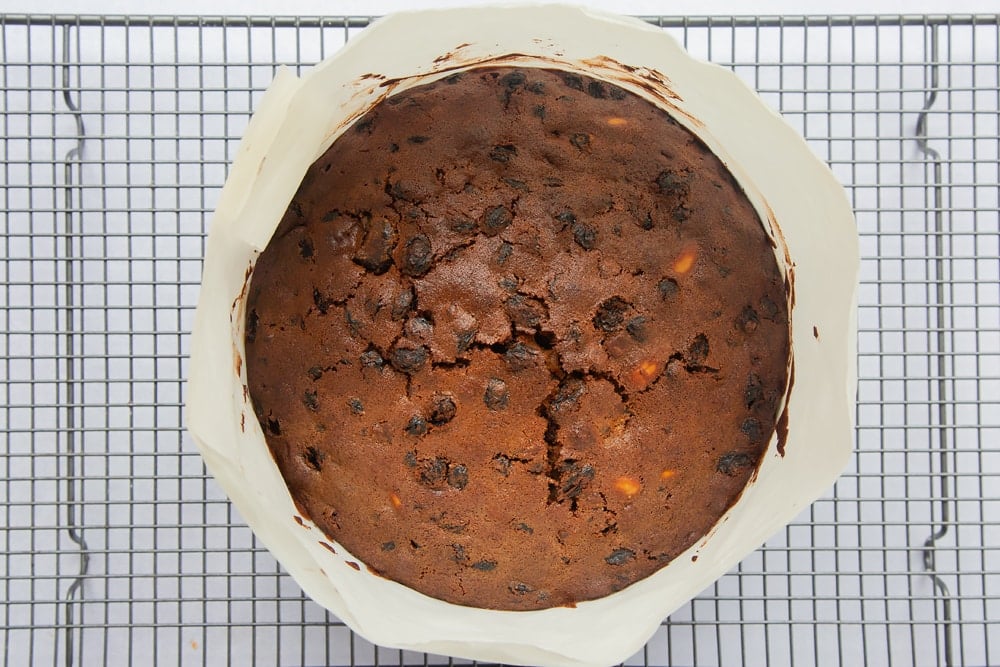 baked christmas fruit cake in a cake tin wrapped in greaseproof paper and brown paper on a wire cooling rack.