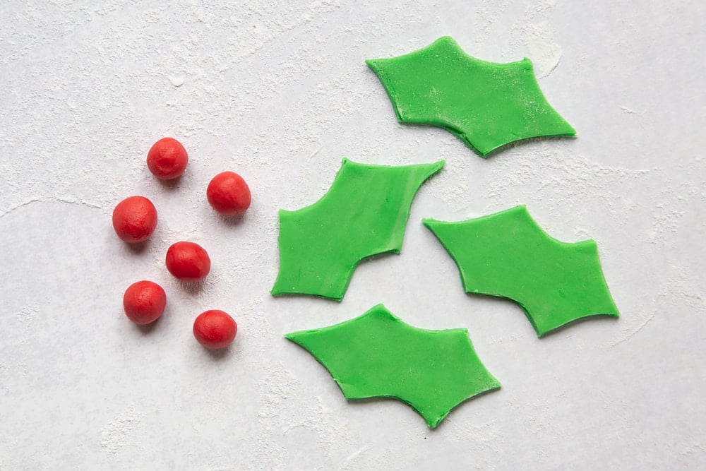 6 small red balls of sugar paste and 4 green holly leaves of sugar paste on a floured surface.