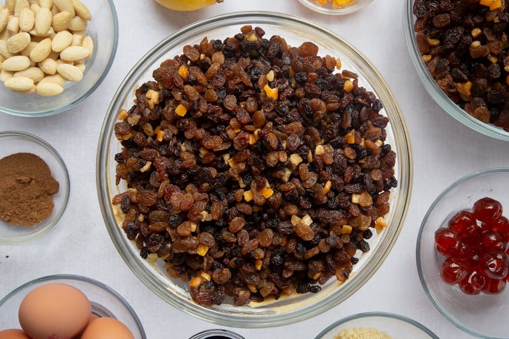 christmas fruit cake base batter mixed together in a large clear bowl topped with chopped dried fruit surrounded by ingredients in smaller clear bowls.