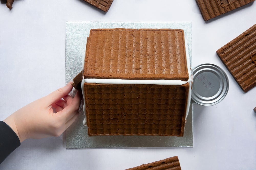 A silver cake board with a gingerbread house on top. A hand positions the gingerbread door on the house.
