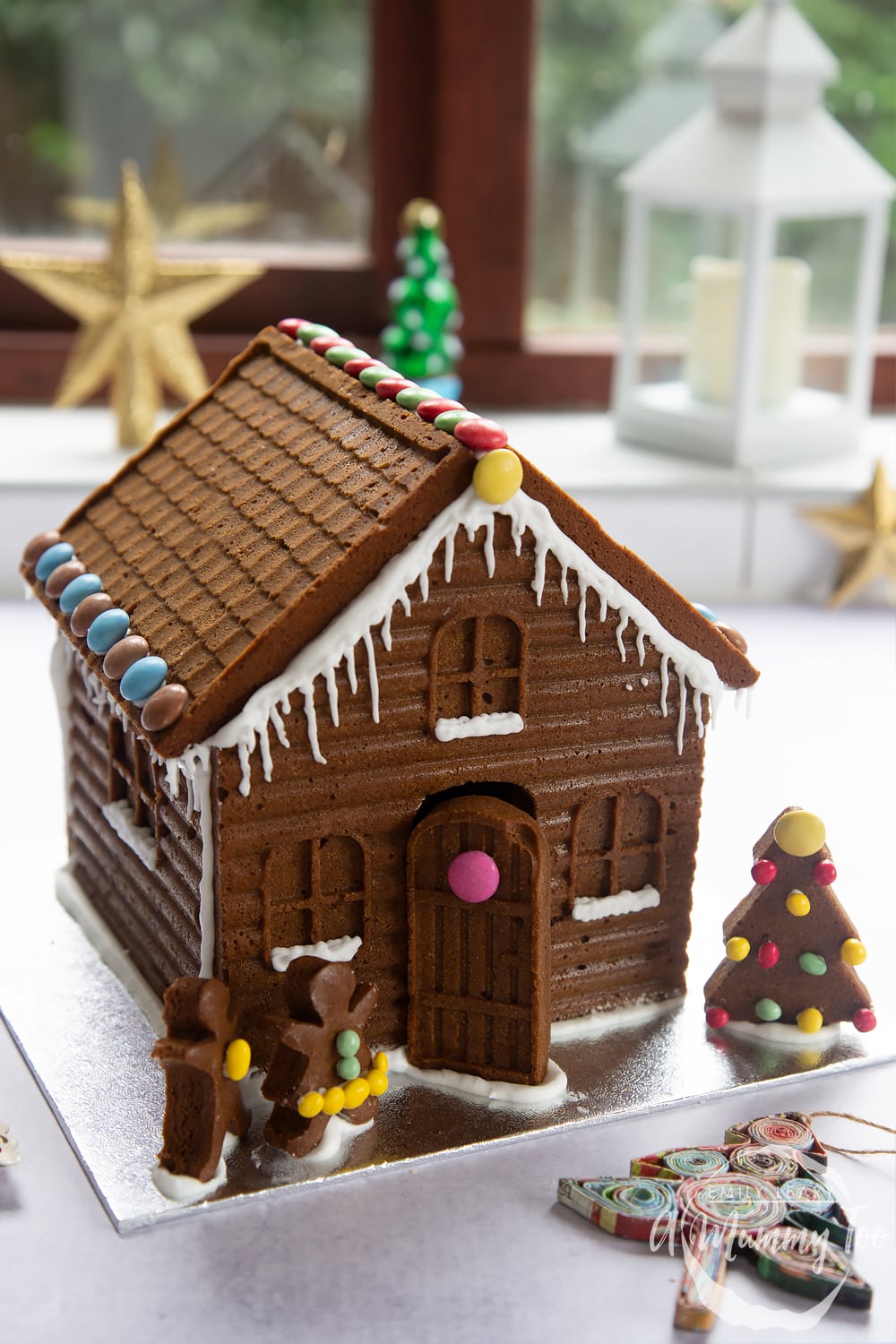A detailed gingerbread house on a silver board. The house is decorated with smarties and mini chocolate beans, as well as royal icing to resemble snow and icicles.
