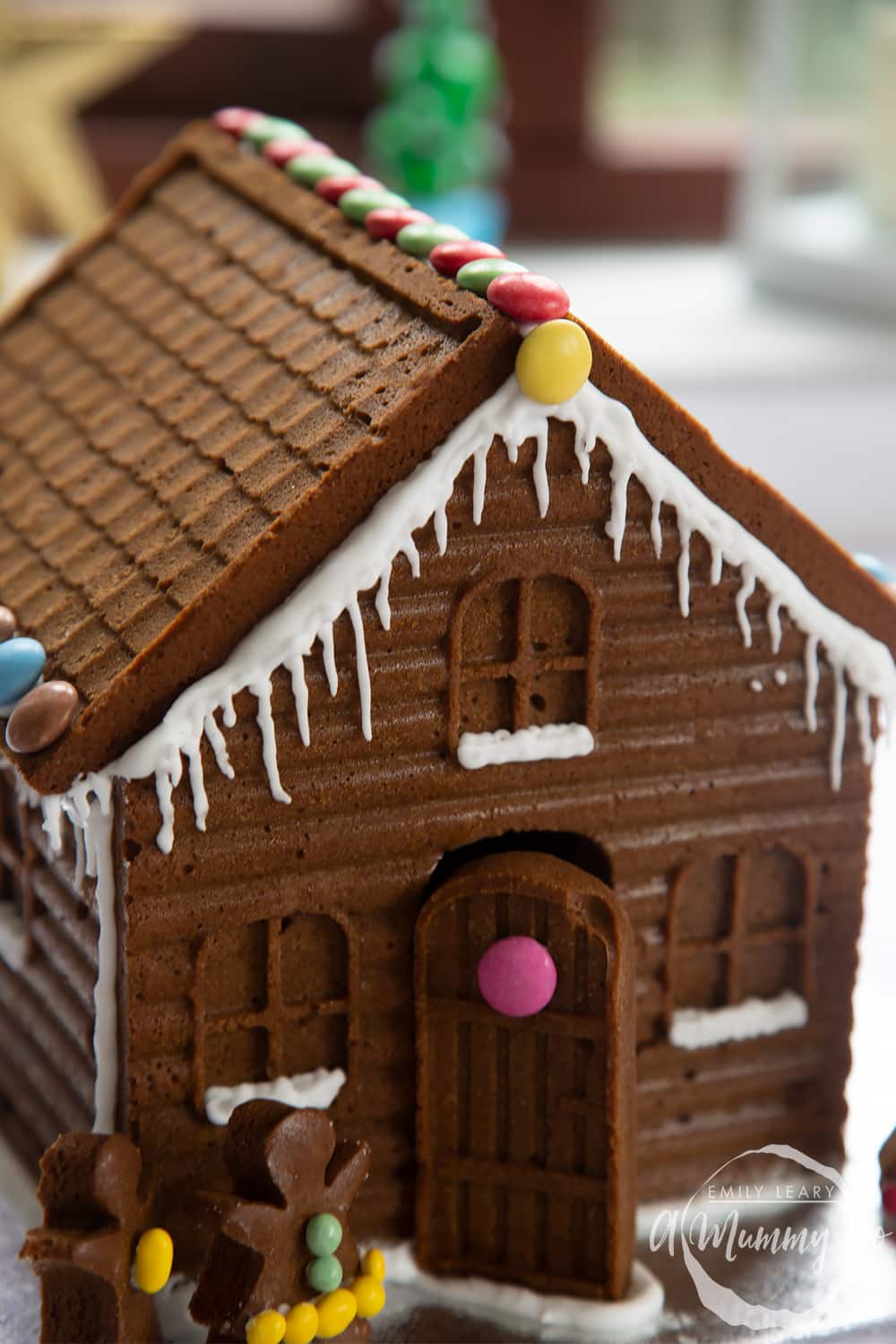 A close up of a detailed gingerbread house on a silver board. Royal icing has been piped along the edges of the roof to resemble snow and icicles.