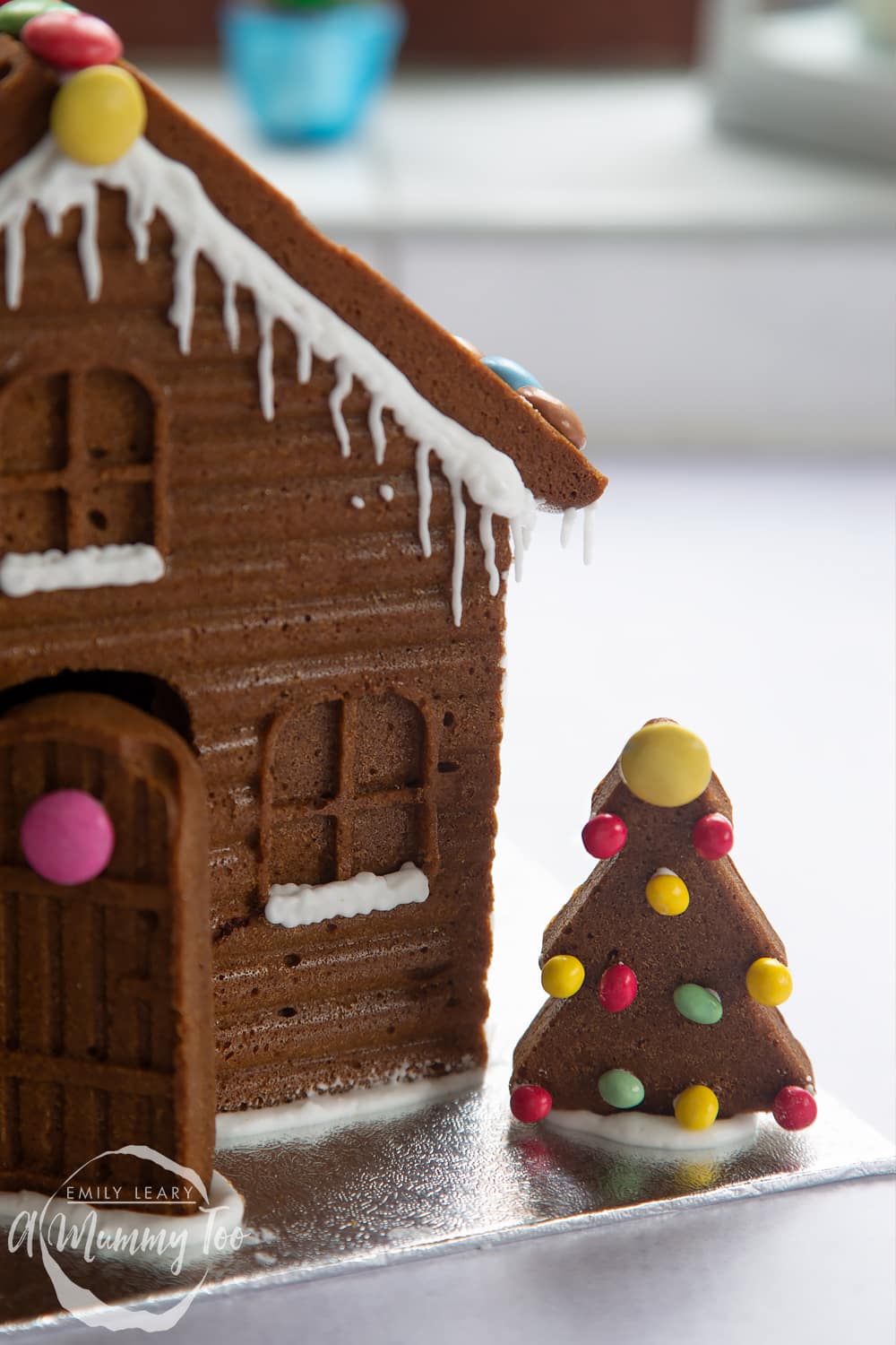 A gingerbread house in close up. The focus is on a gingerbread Christmas tree, decorated with mini chocolate beans.