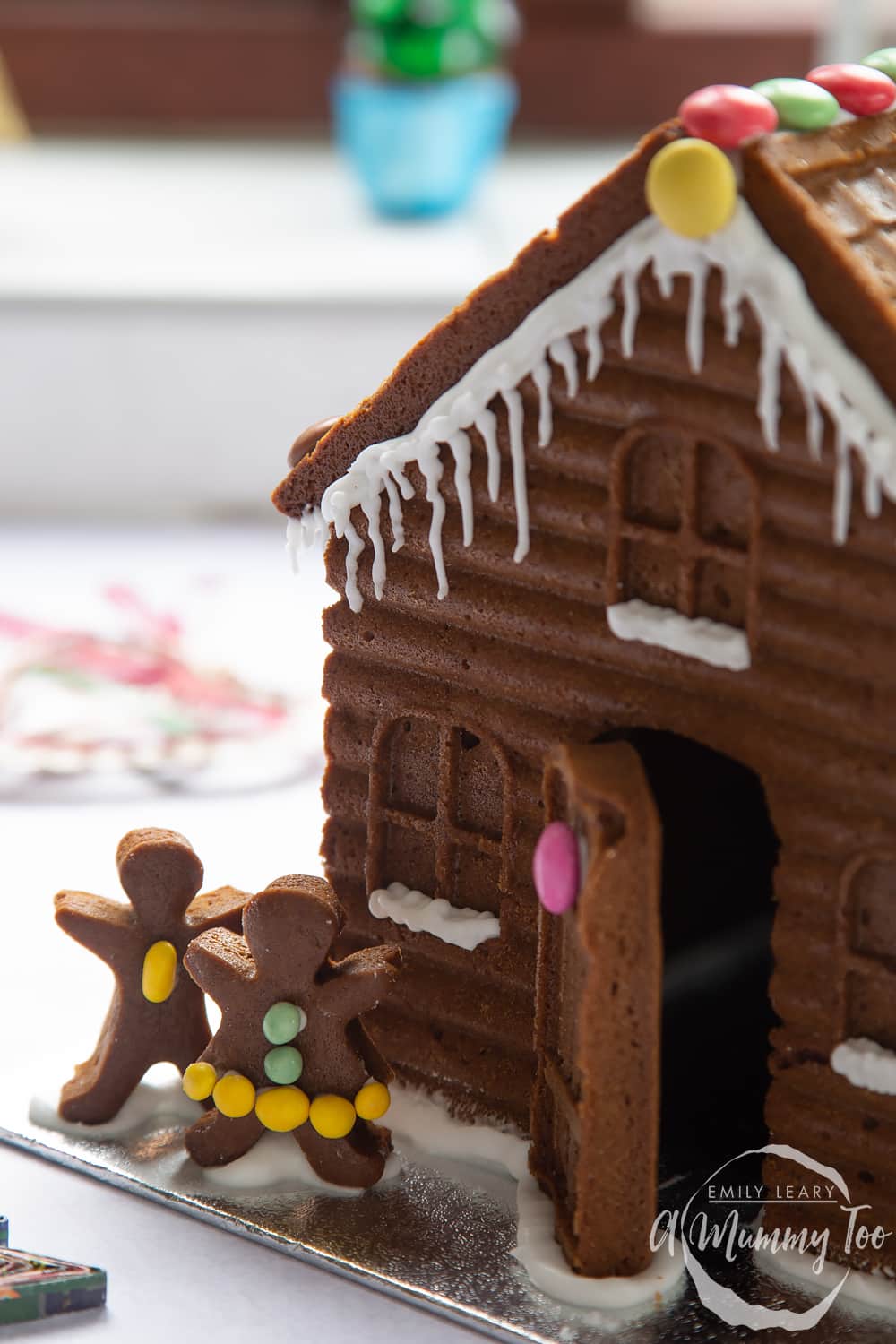 A gingerbread house in close up. The focus is on two gingerbread people, decorated with mini chocolate beans.