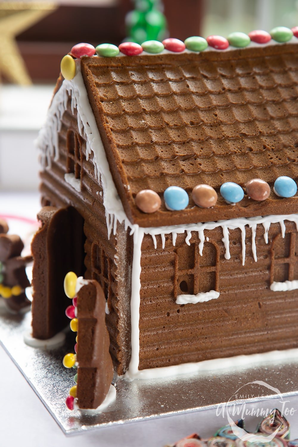 A gingerbread house stands on a silver board. The sills of the windows have been piped with royal icing to resemble snow. 