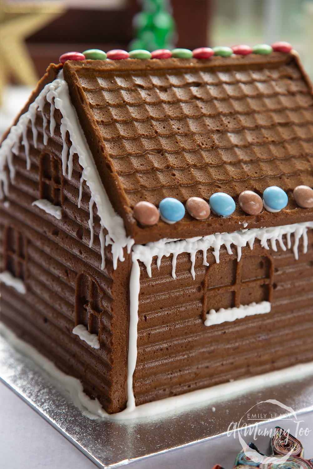 A gingerbread house stands on a silver board. The sills of the windows have been piped with royal icing to resemble snow, while icing icicles hang from the roof, which is also decorated with chocolate beans.