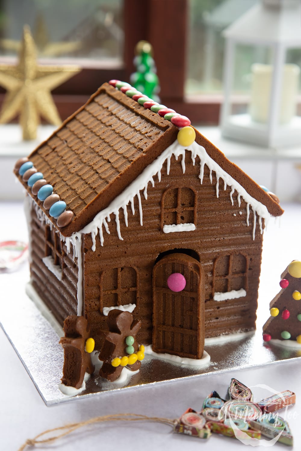 A detailed gingerbread house on a silver board. The house is decorated with smarties and mini chocolate beans. 