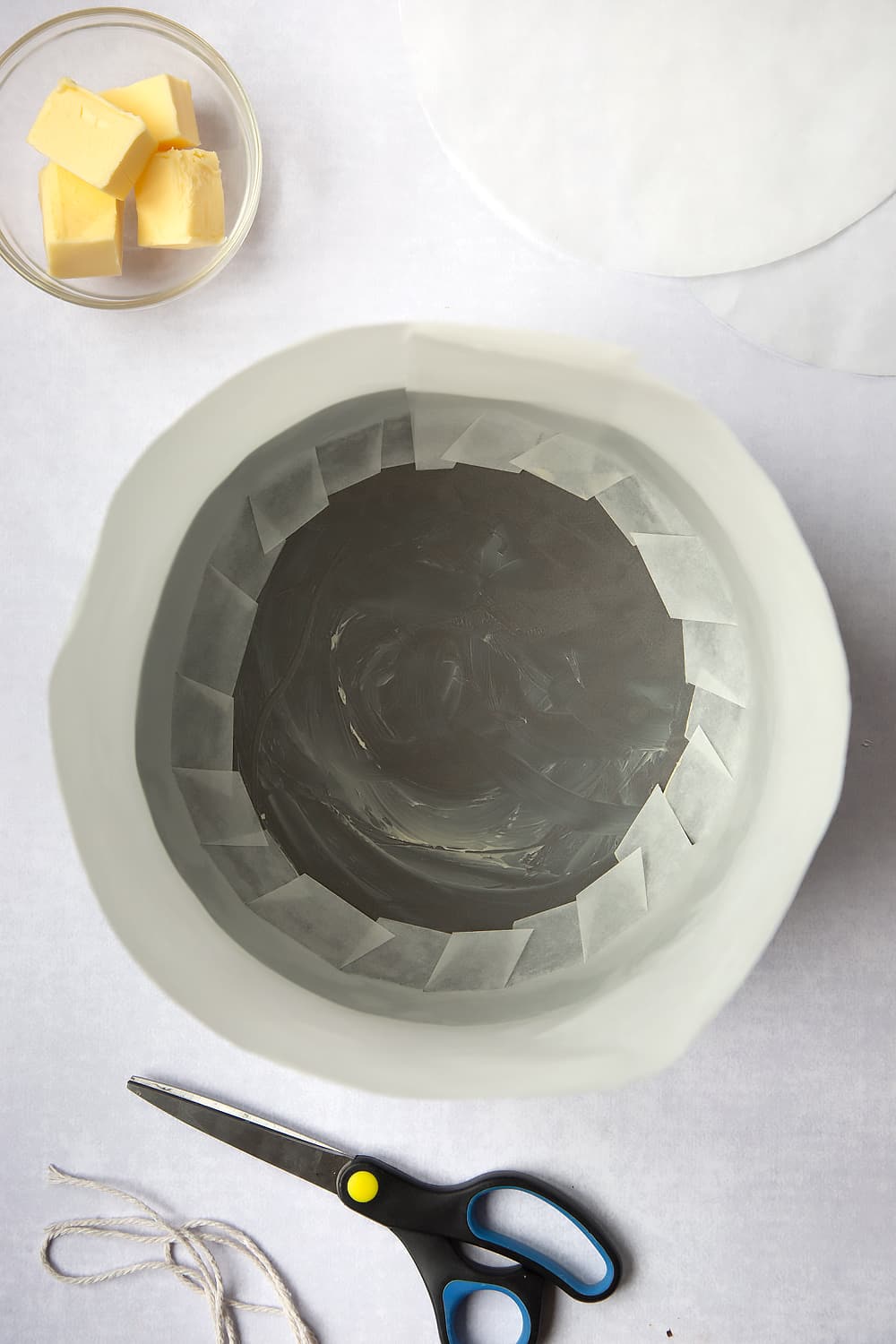 A baking tin which has been greased with butter, with baking paper situated around the outside with the snipped edges of the lip on inside covering some of the base of the cake tin. 
