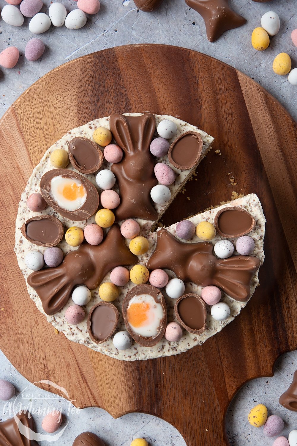 No-bake Easter cheesecake shown from above. The cheesecake is topped with mini chocolate eggs, creme eggs and chocolate bunnies. The cheesecake sits on a wooden board and has a slice taken from it.