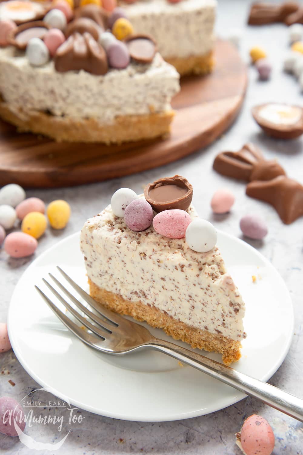 A slice of no-bake Easter cheesecake on a white plate with a fork beside it. The cheesecake is topped with mini chocolate eggs. More cheesecake is visible in the background. 