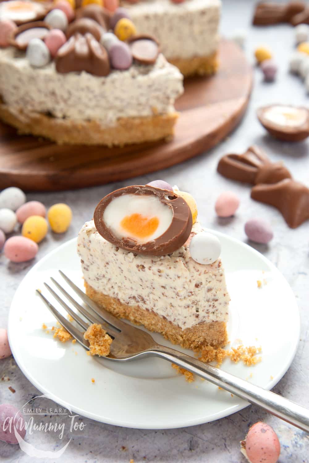 A slice of no-bake Easter cheesecake on a white plate with a fork beside it. The cheesecake is topped with mini chocolate eggs and half a creme egg. More cheesecake is visible in the background. A piece has been taken from the slice and crumbs site around and on the fork.