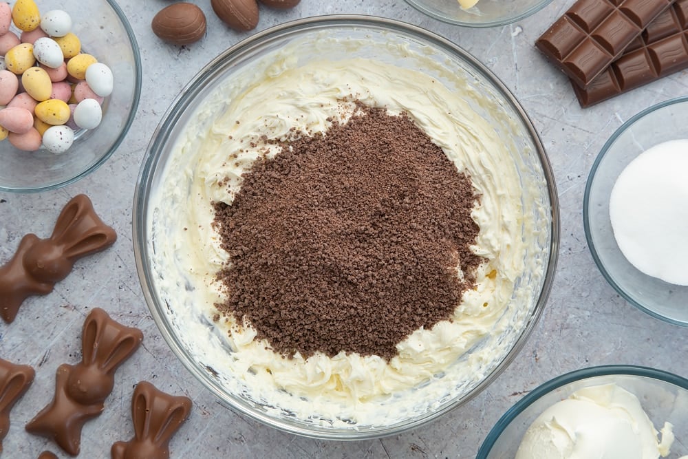 A glass bowl containing double cream, cream cheese and sugar beaten together to a stiff mix. Grated chocolate is poured on top. Around the bowl are ingredients to make no-bake Easter cheesecake.