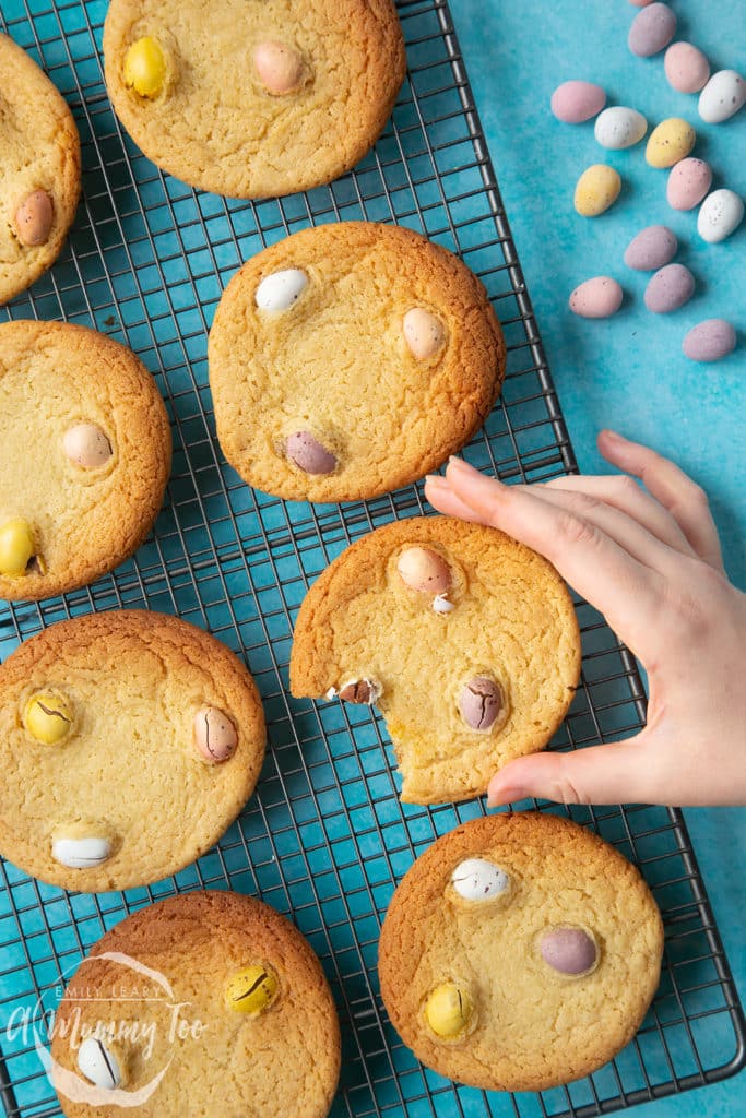 An overhead view of cookies cooling on a tray. A childs hand is reaching in to pick up one cookie with a bite out of it