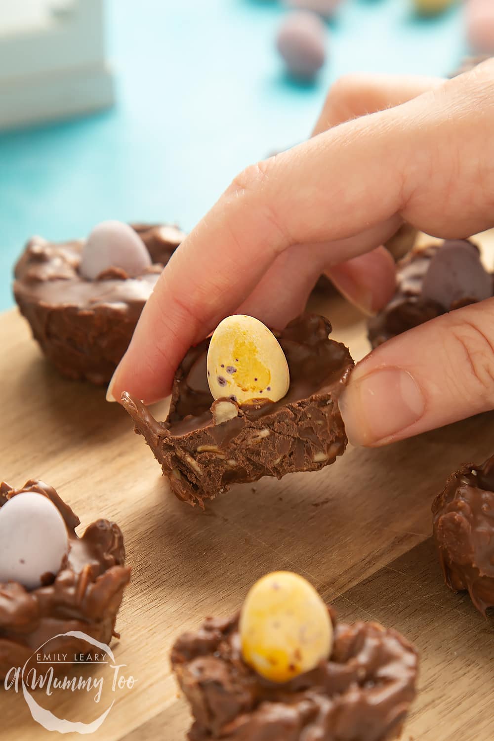 Front angle shot of crunchy muesli chocolate nest in a hand, served on a wooden board with a mummy too logo in the lower-left corner