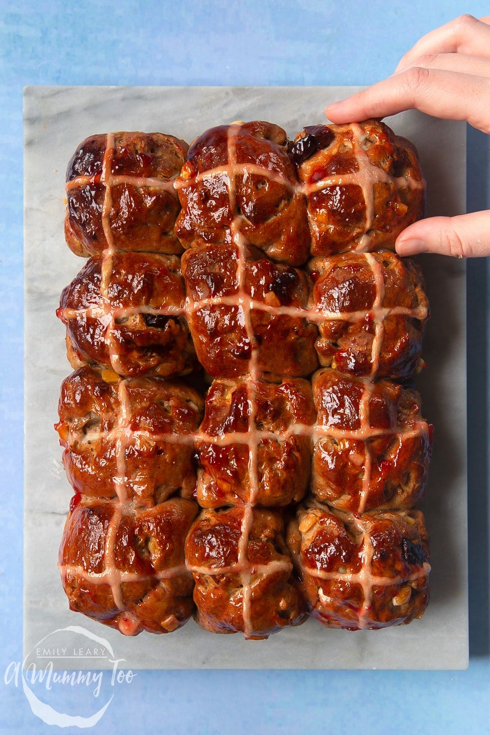 12 vegan hot cross buns on a marble board. A hand reaches in as if to take one.