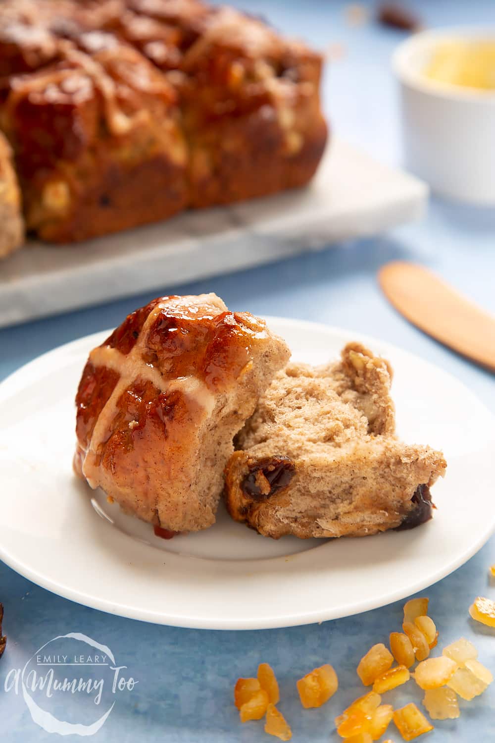 A vegan hot cross bun cut open on a white plate. More vegan hot cross buns are shown on a marble board in the background.