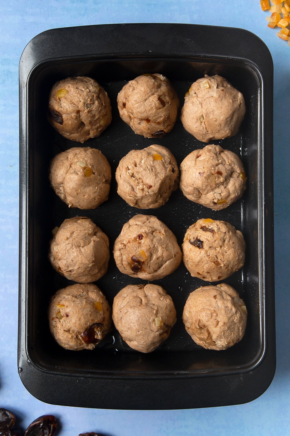 Vegan hot cross buns dough with mixed peel and chopped dates, divided into 12 balls and placed in an oiled tray.