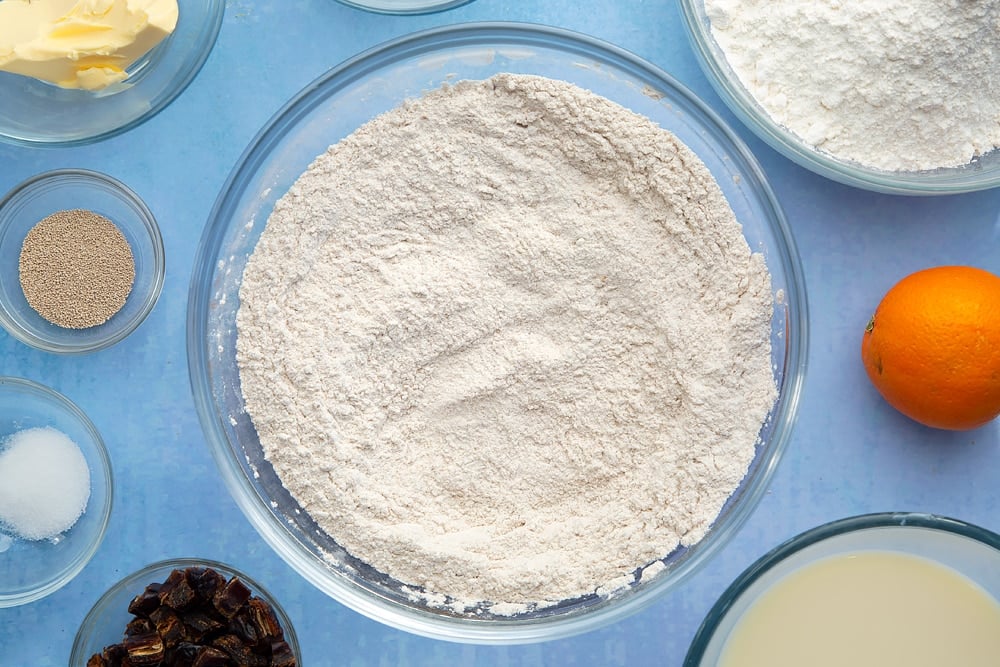 A bowl containing flour, yeast, sugar, salt, cinnamon, ginger and nutmeg, whisked together. The bowl is surrounded by the ingredients to make vegan hot cross buns.