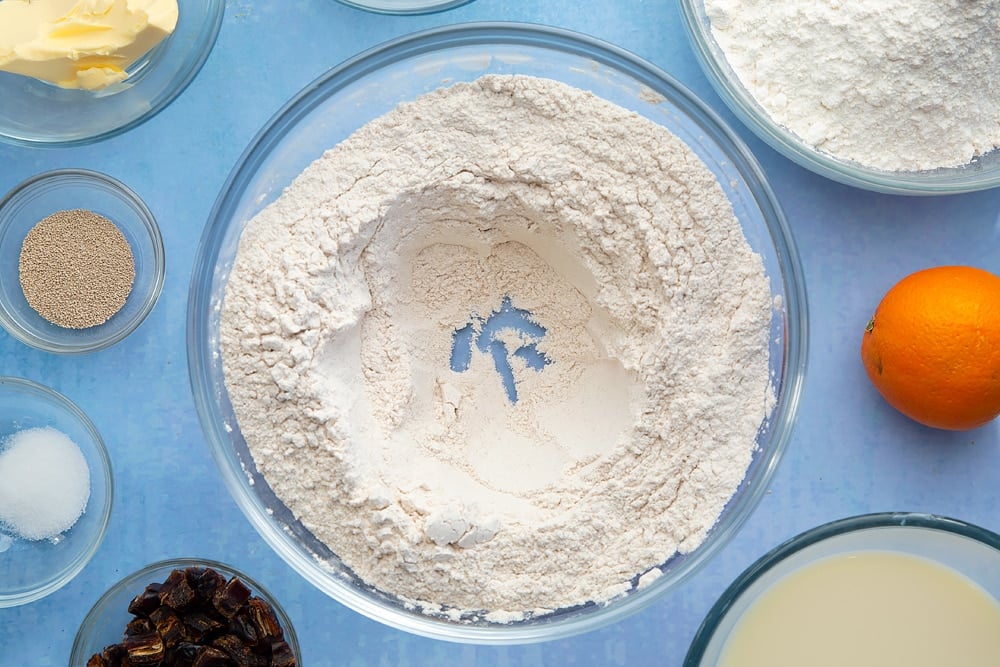 A bowl containing flour, yeast, sugar, salt, cinnamon, ginger and nutmeg, whisked together, with a well made in the centre. The bowl is surrounded by the ingredients to make vegan hot cross buns.