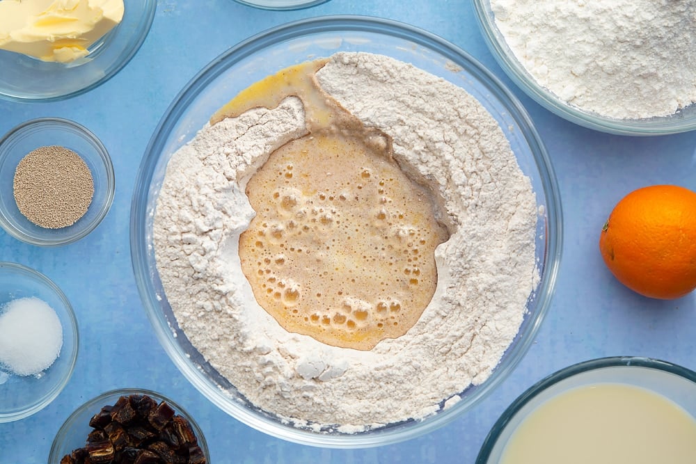 A bowl containing flour, yeast, sugar, salt, cinnamon, ginger and nutmeg, whisked together, warm milk and margarine is poured into the well in the centre. The bowl is surrounded by the ingredients to make vegan hot cross buns.