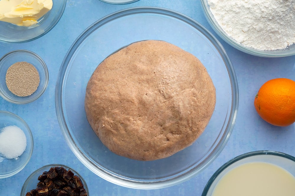 A bowl containing a smooth ball of vegan hot cross buns dough that has just proved. The bowl is surrounded by vegan hot cross bun ingredients.