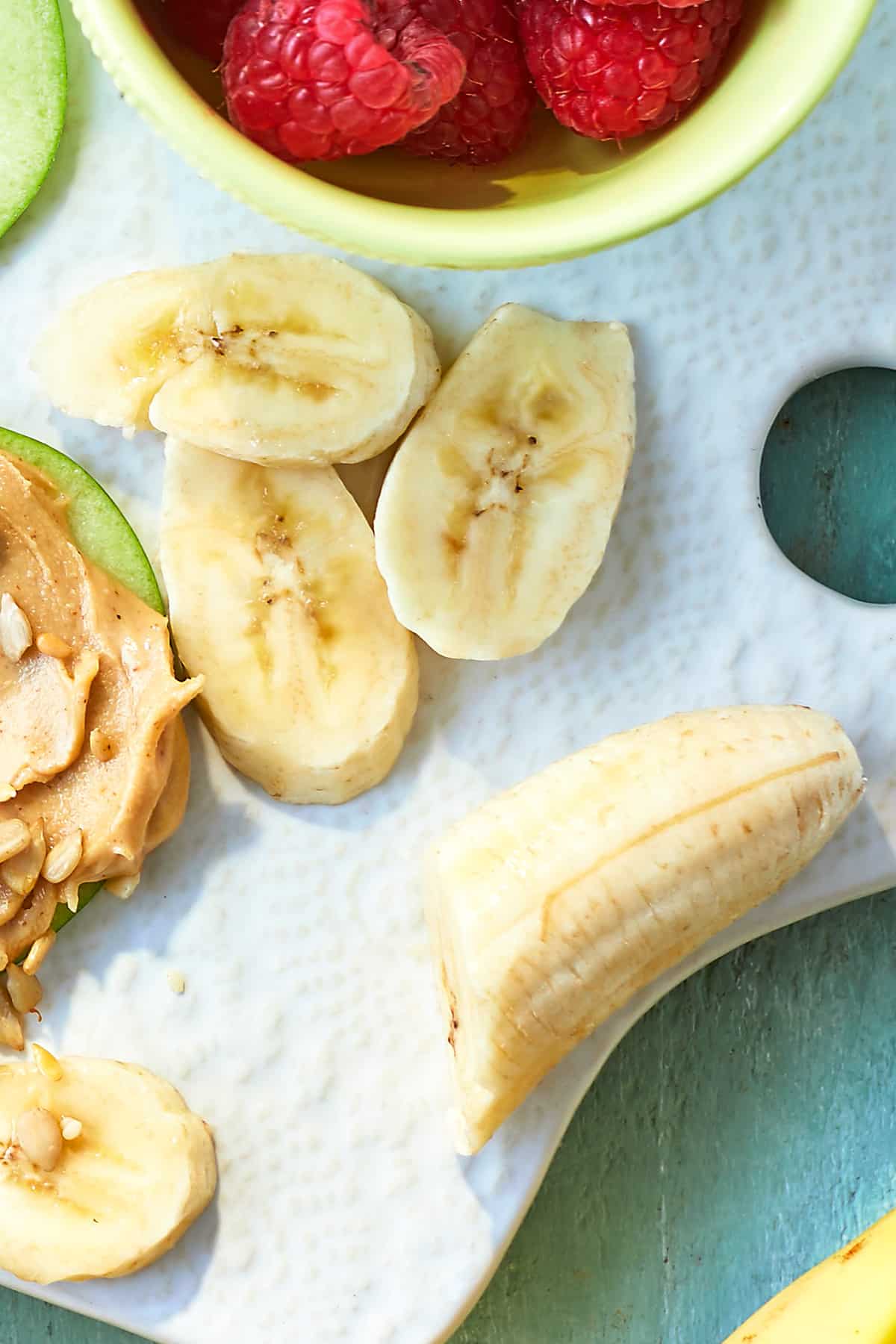 Sliced banana on a white board, surrounded by ingredients for apple slices with peanut butter.