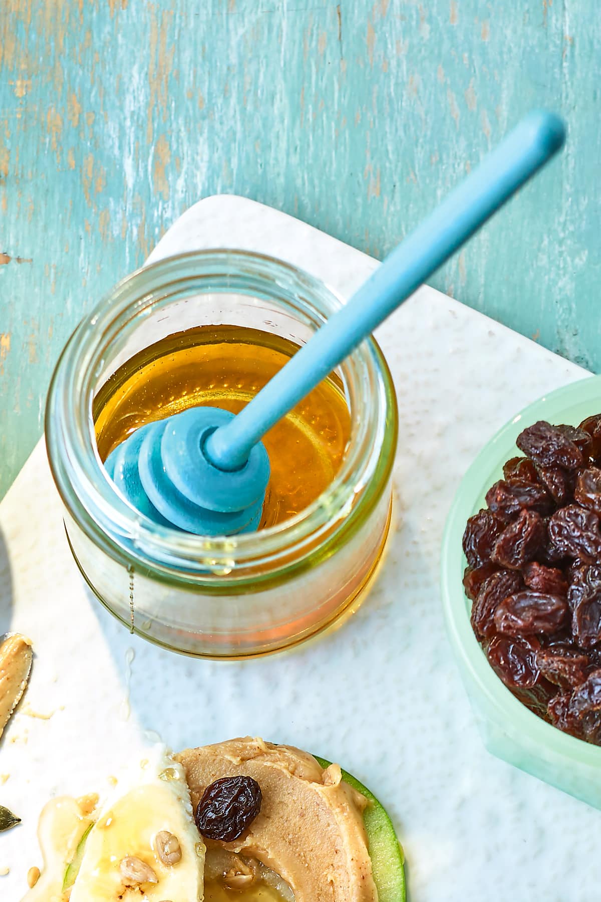 Honey in a jar on a white board. Also show are ingredients for apple slices with peanut butter.