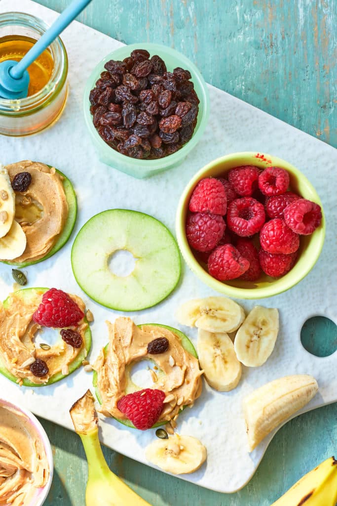 Overhead view of doughnut shaped apple slices and bowls of fruit. All but one of the apple slices are covered with peanut butter and more fruit.